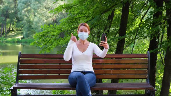 Young Woman Sits Alone in a Park in a Mask and Talks Over a Videocall During the Epidemic 