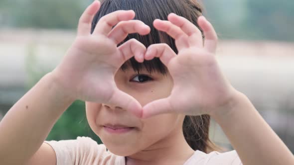 Cute Asian preschooler smiling happily making heart-shaped hands on eyes on green nature.
