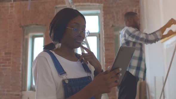 Young African Woman Using Tablet Pc While Boyfriend Using Spirit Level Measuring Wall on Background