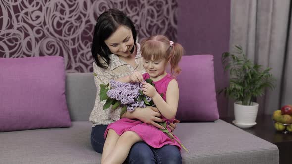 Little Daughter and Mother Sitting on Sofe with Bouquet of Lilac Purple Flowers