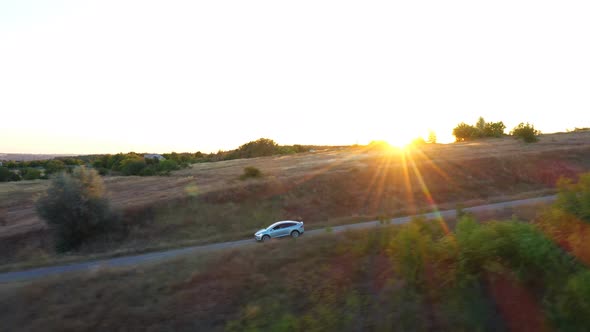 Aerial Shot of Electrical Car Driving on Country Road with Bright Sunset at Background