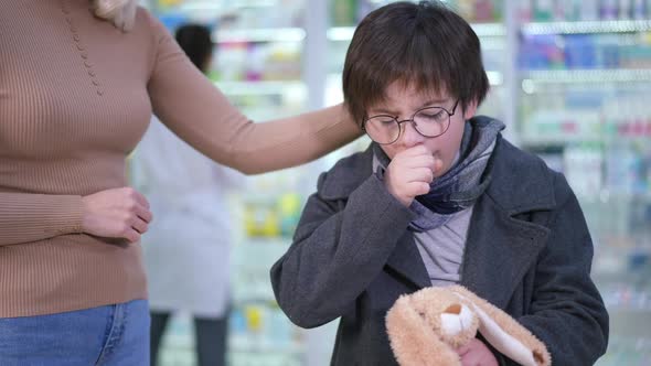 Unwell Caucasian Boy Coughing Standing in Pharmacy with Unrecognizable Woman Caressing Hair
