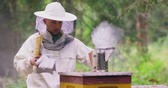 Young Bearded Male Beekeeper in White Protective Suit Approaches to the Hive and Puts the Smoking