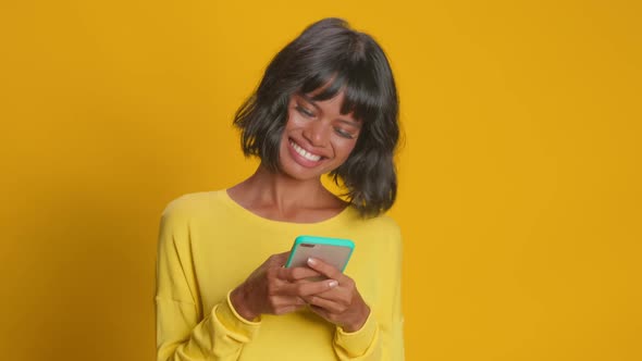 Young Happy Dark Skinned Woman Checks Message on Smartphone Against Orange Wall