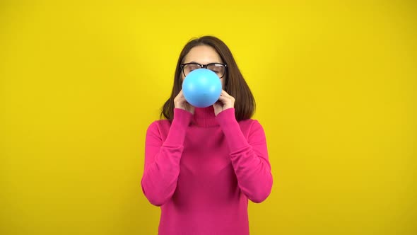 A Young Woman Inflates a Blue Balloon with Her Mouth on a Yellow Background. Girl in a Pink