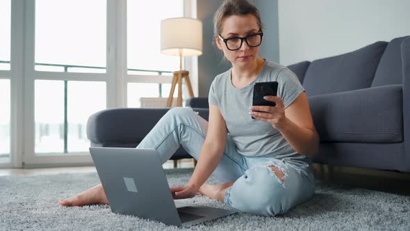 Casually Dressed Woman Sitting on Carpet with Laptop and Smartphone and Working in Cozy Room