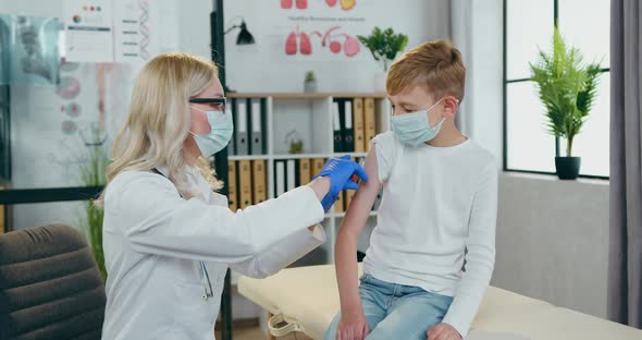 Female Doctor in Medical Mask Doing Injection in Boy's Arm which Sitting on the Reviewing Table