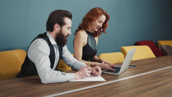 Woman Typing Text on a Laptop in the Office and Next To a Man with a Beard Writing Text