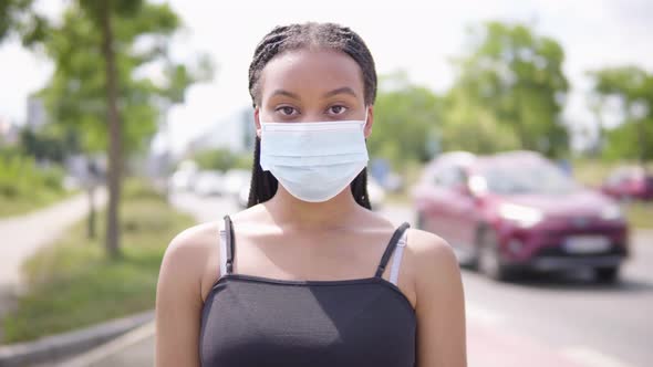 A Young Black Woman Puts on a Face Mask and Looks at the Camera - a Busy Road in an Urban Area