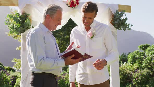 Smiling caucasian senior male wedding officiant holding book and groom standing in outdoor altar