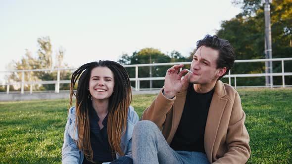 Happy Young Hipster Couple Sitting on Grass and Smoking Marijuana Joint in Park Slow Motion