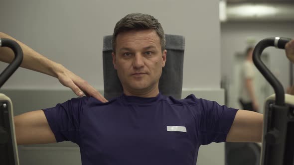 Middle-aged Sportsman Doing Strength Exercises on the Simulator for Hands Looking at Camera