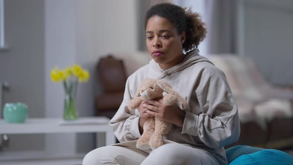 Portrait of Depressed Young African American Woman Hugging Toy Rabbit Sitting at Home Indoors