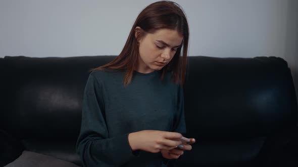 The Portrait of a Freckled Redhaired Woman Playing Focused on the Phone Sitting on the Black Sofa at