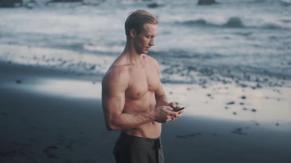 Topless Young Sportsman Uses His Smartphone at the Sea Shore