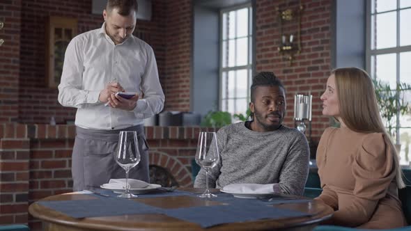 Confident Wealthy Interracial Young Couple Ordering Dinner in Luxurious Restaurant As Waiter Writing