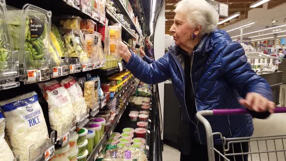 Elderly woman looking at cut squash in plastic container in grocery store.