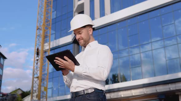 Architect Engineer in a White Shirt and Helmet on a Background of a Modern Glass Building Works in a