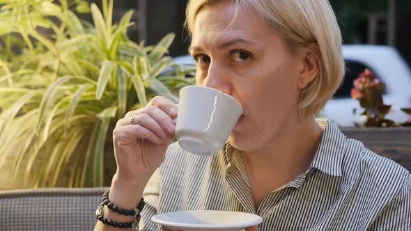 Portrait A Woman Drinks Coffee Holding a White Cup in Her Hands While Sitting in a Summer Street