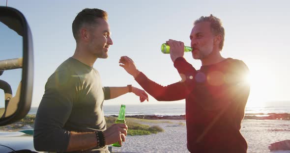 Happy caucasian gay male couple standing by car drinking bottles of beer on sunny day at the beach