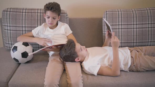 Caucasian Boy Sitting at Couch with Notebook and Ball, His Twin Brother Laying on His Knees