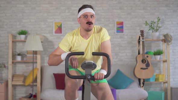 Funny Tired Athlete From the 80's with a Mustache Engaged at Home on a Exercise Bike Slow Mo