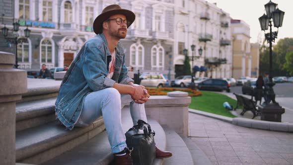 Young Stylish Handsome Redbeard Man with Bag and in Hat Sitting on Stairs in Old City Center Slow