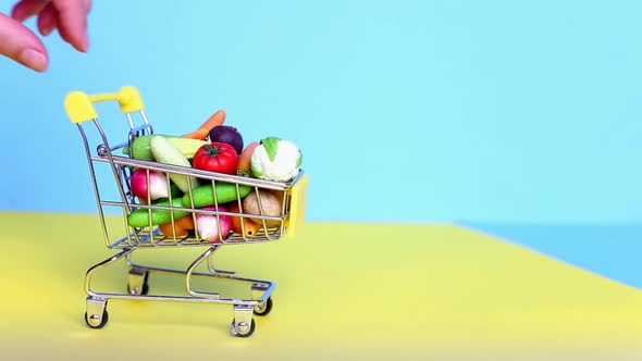 hand holding shopping cart with fruits and vegetables on green and yellow background.