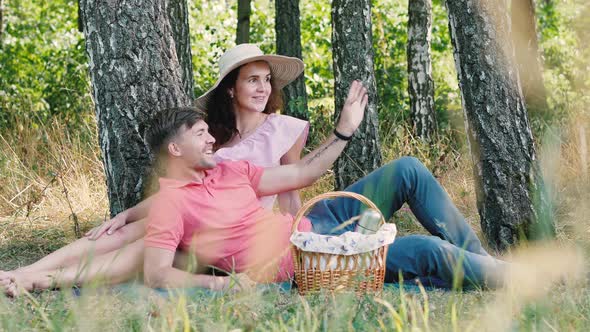 Picnic Time. Happy Young Couple Sitting and Smiling Outdoors. Guy Points His Hand To Friends and