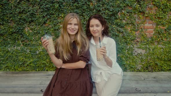 Two Women Sit on a Bench Near a Wall of Vines and Drink Drinks