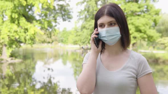 A Young Caucasian Woman in a Face Mask Talks on a Smartphone in a Park