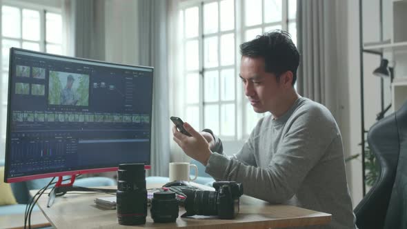 Asian Video Editor Man Using Mobile Phone While Using Desktop Computer For Editing Video At Home