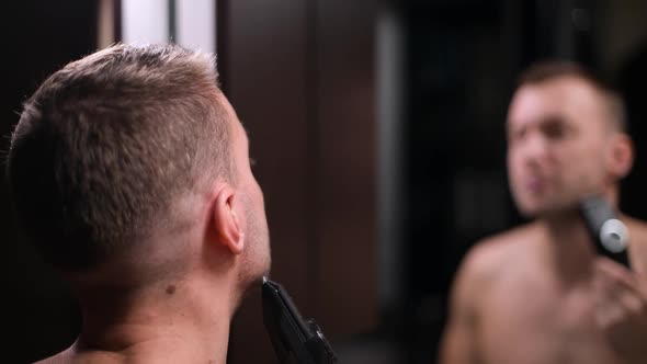 a man cutting his beard with a trimmer in front of a mirror