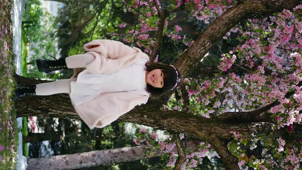 Korean Girl in a White Light Fur Coat and a Headband Stands in a Garden with Cherry Blossoms