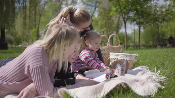 Two Mothers on the Blanket in the Park with Their Children. Teenage Blond Boy Lying and Reading the