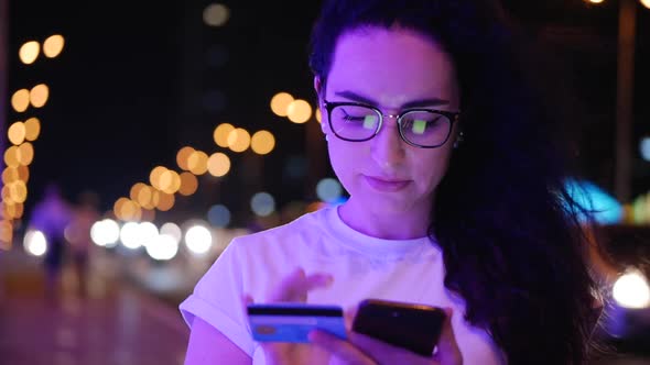 Pretty Woman Stands on the Street in a White T-shirt and Glasses, Makes Online Payments By Credit