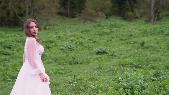 A Young Beautiful Woman in a White Wedding Dress Stands in a Field with Horses