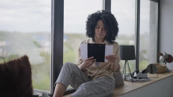 Wide Shot Portrait of Serious Woman Sitting on Windowsill Analyzing Business Project