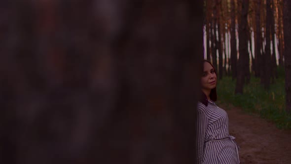 Young Woman Stands Leaning on Tree on Walk in Forest