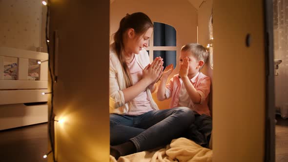 Smiling Cheerful Boy Playing Patty Cake with Young Mother in Toy Small House at Night