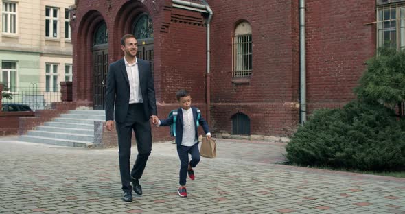 Little Child with Backpack Hopping and Smiling While Going with Father. Cheerful Man in Suit Holding