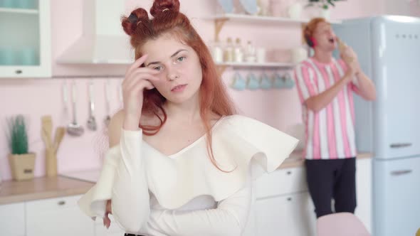 Annoyed Redhead Woman Looking at Camera As Blurred Man in Headphones Singing at the Background Using