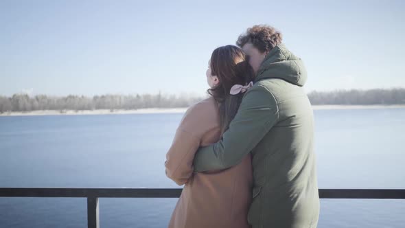 Back View of Young Caucasian Man Kissing Girlfriend on River Bank and Smiling. Happy Married Couple