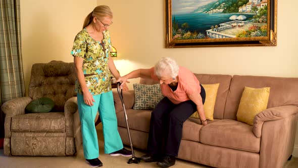 Home healthcare nurse helps elderly woman get up from couch and use a cane.