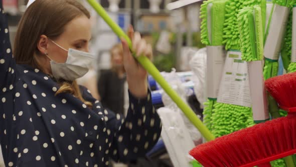 Young Woman in a Protective Mask Choosing a Mop in a Hardware Store