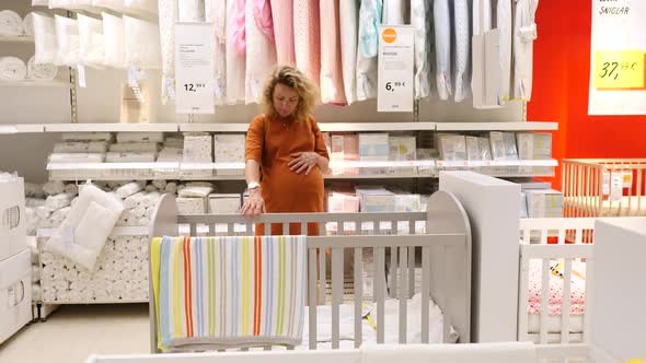 Young Pregnant Woman Choosing Cot Or Baby Bed In Furniture Store