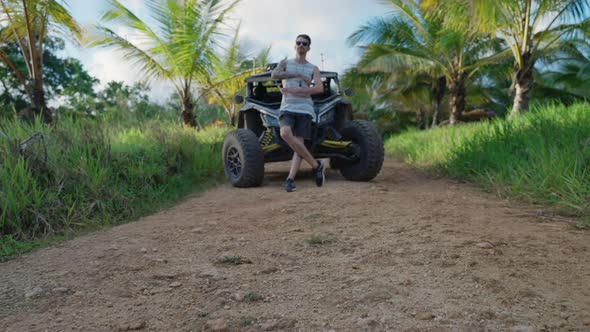 Male Wearing Sunglasses Leaning Against Bonnet Of Dirt Buggy In Empty Path In Punta Cana. Low Angle,