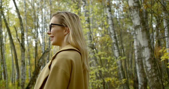 Close-up Portrait of a Young Smiling Blonde Girl in Glasses and a Coat, She Walks Along a Path
