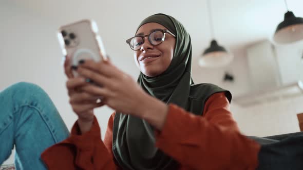 Happy Muslim woman wearing eyeglasses texting by phone