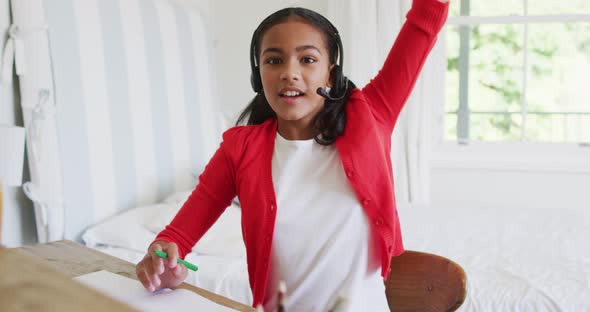 Mixed race girl at home in online school class, using headset raising hand